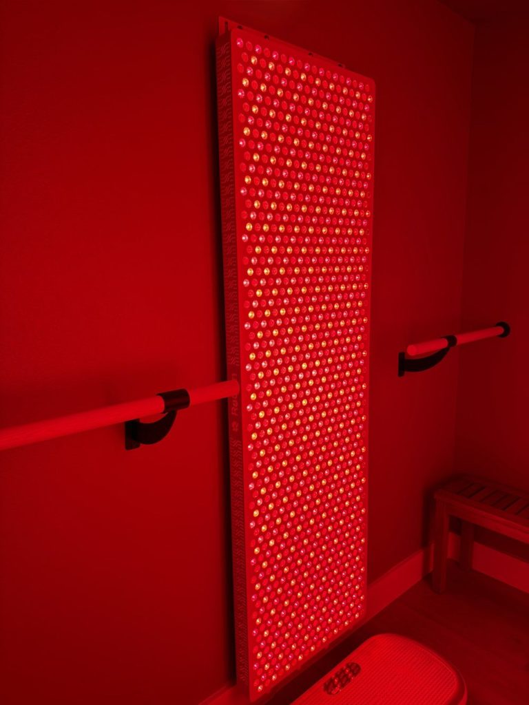 Full-length red light therapy panel glowing against the red wall at Adoration